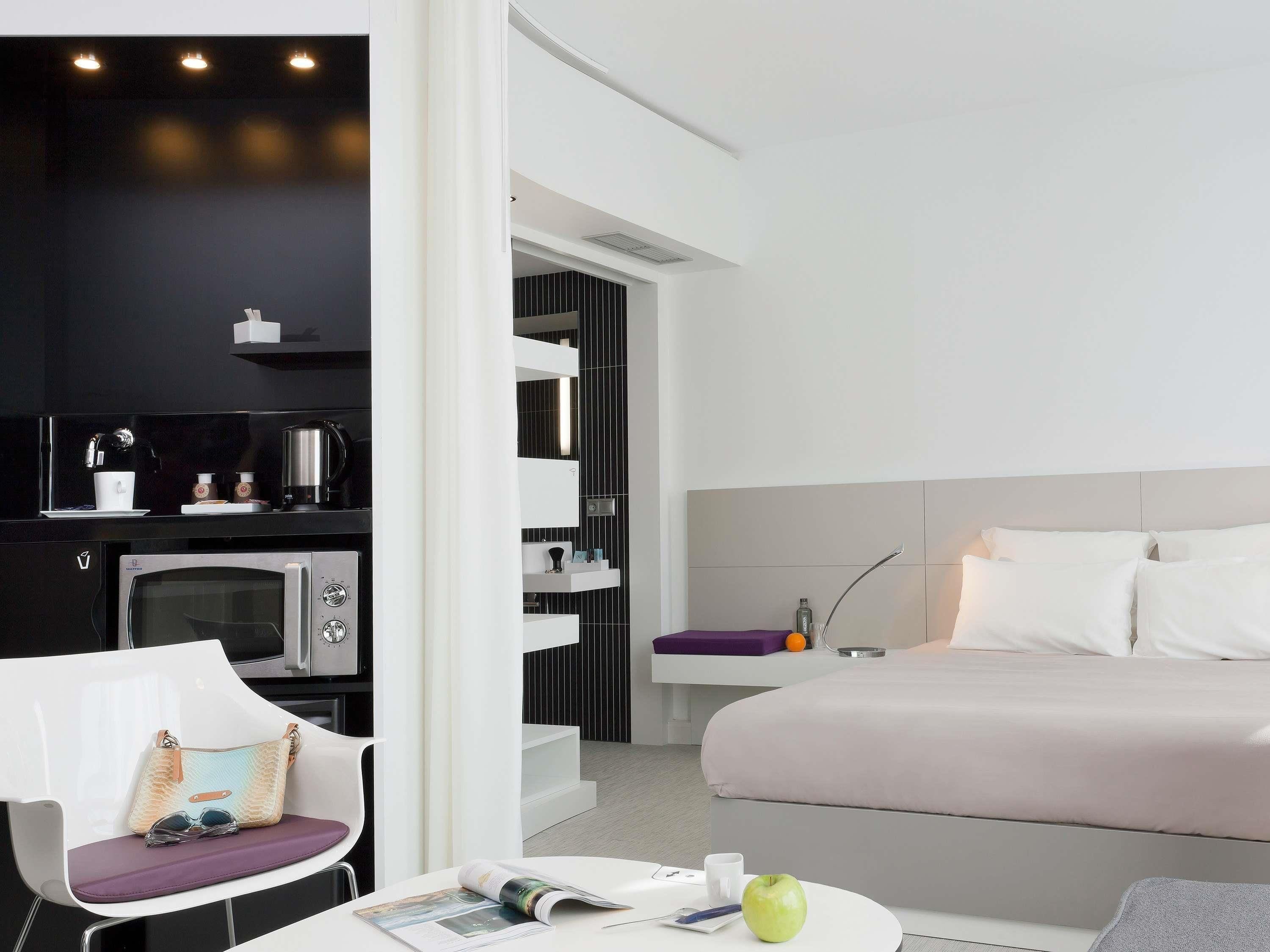 A modern hotel room with a bed, a seating area with a magazine and a cup, a kitchenette with a coffee machine, and a bathroom visible through an open door.