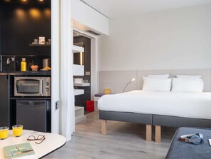 Ein modernes Hotelzimmer mit Bett, einer kleinen Küchenzeile, einem Esstisch und einem Badezimmer.