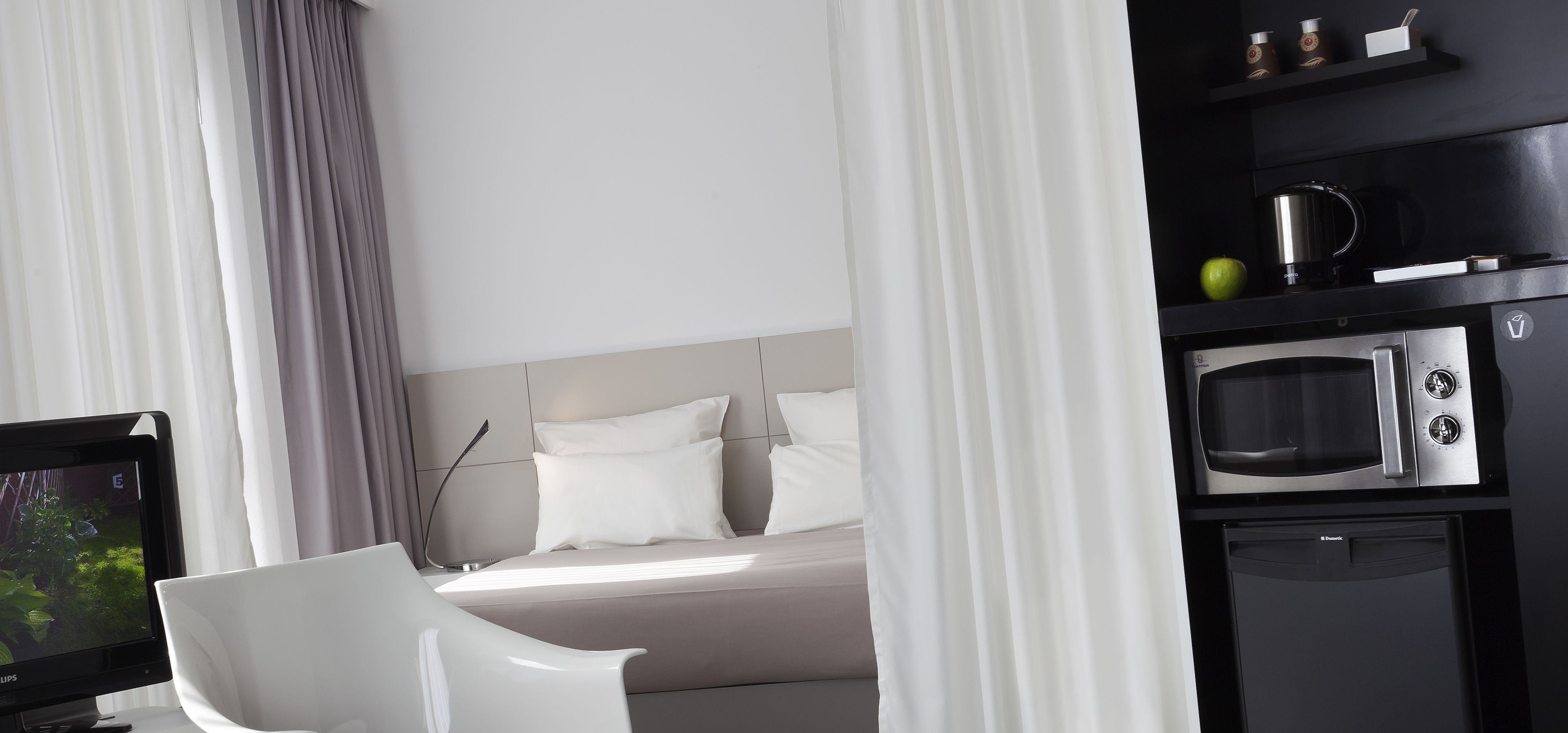 Ein modernes Hotelzimmer mit einem Bett, einem weißen Stuhl, einem Fernseher und einer Küchenzeile mit einer Mikrowelle.