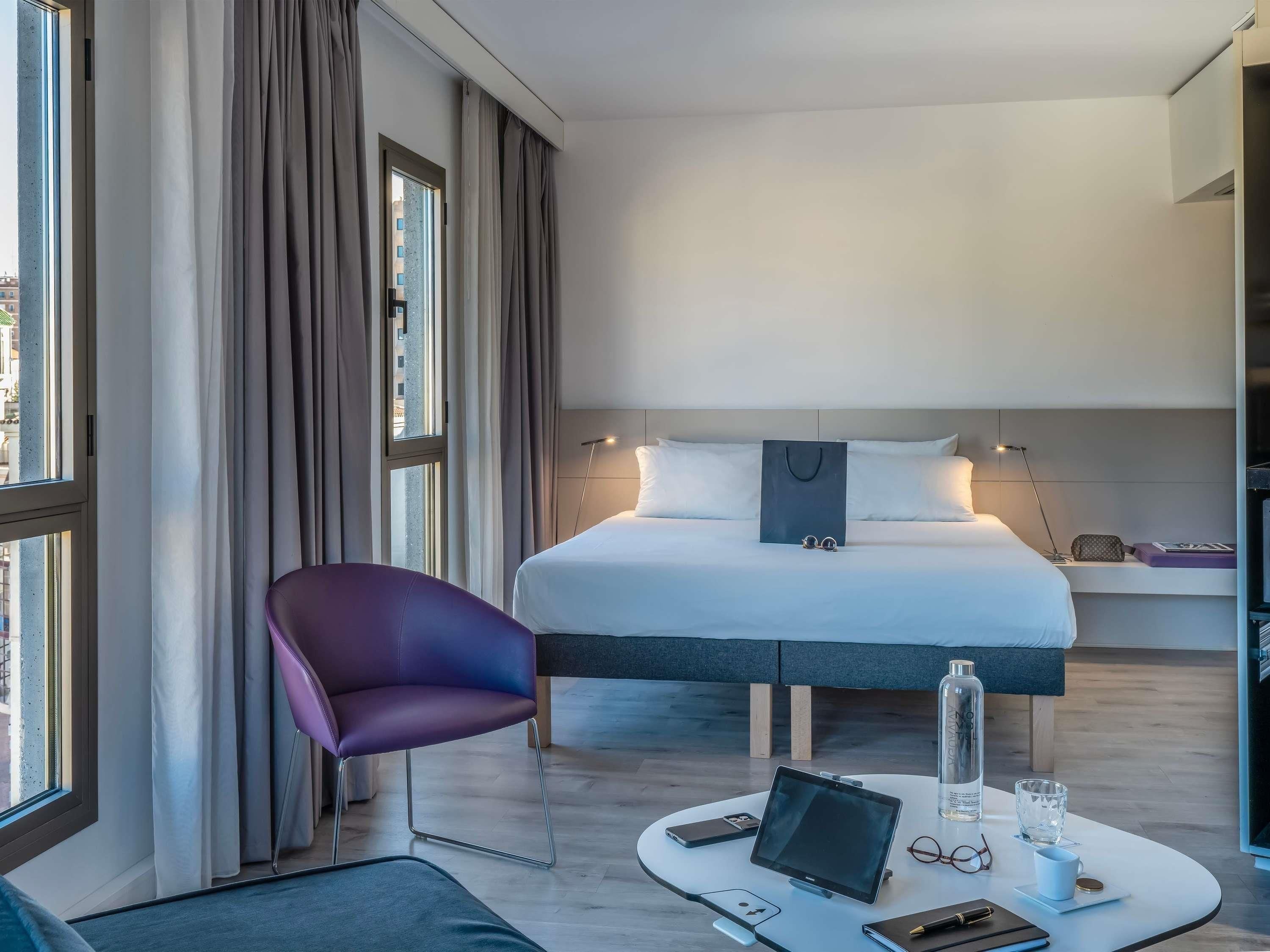 Een moderne hotelkamer met een bed, een paarse stoel, een ronde tafel met een laptop en een nachtkastje met een lamp en een glas.