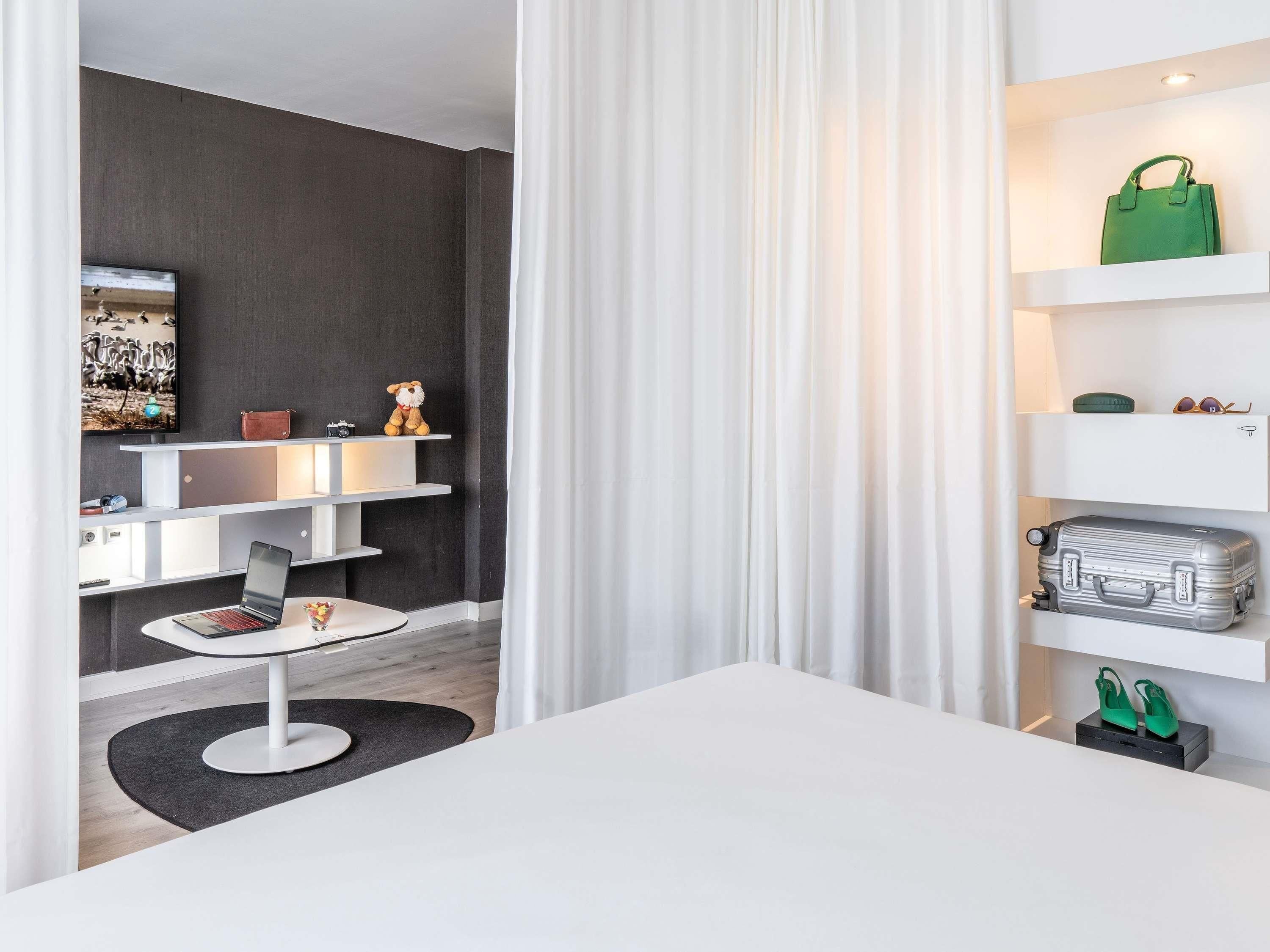 Una habitación de hotel moderna con un escritorio blanco, una computadora portátil, un osito de peluche y un televisor sobre una pared oscura.