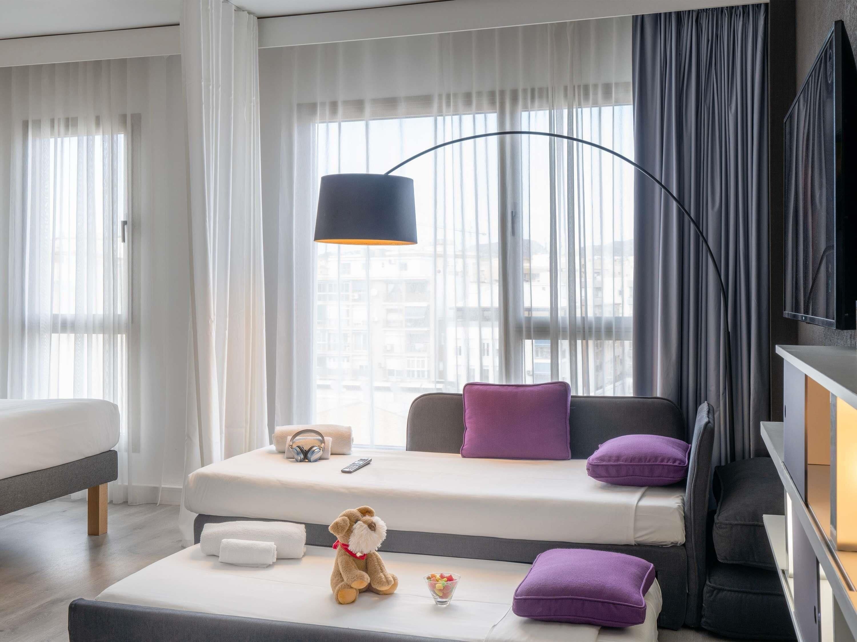 A modern hotel room with a sofa bed, a flat-screen TV, a lamp, and a teddy bear on a small table.