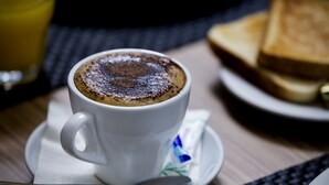 A cup of coffee with a frothy top, a spoon, and a plate with toast on a table. - Novotel Suites Malaga Centro (Málaga)