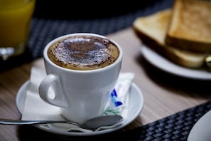 A cup of coffee with a frothy top, a spoon, and a plate with toast on a table. - Novotel Suites Malaga Centro (Málaga)