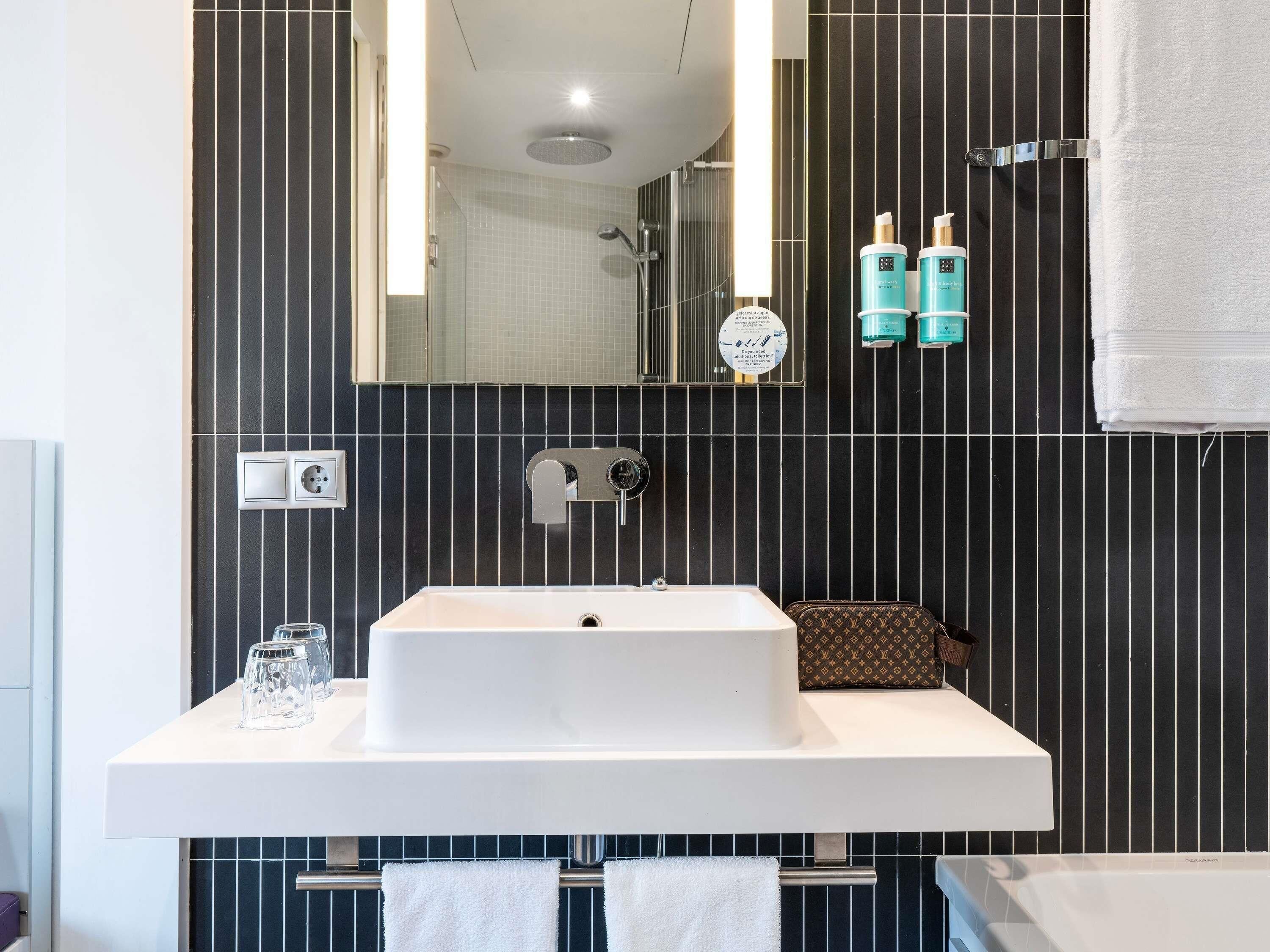 Een moderne badkamer met een grote spiegel, een witte wastafel en een douche. Er staan twee flessen shampoo en een handdoekrek met handdoeken.