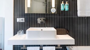 A modern bathroom with a large mirror, a white sink, and a shower. There are two bottles of shampoo and a towel rack with towels. - Novotel Suites Malaga Centro (Málaga)