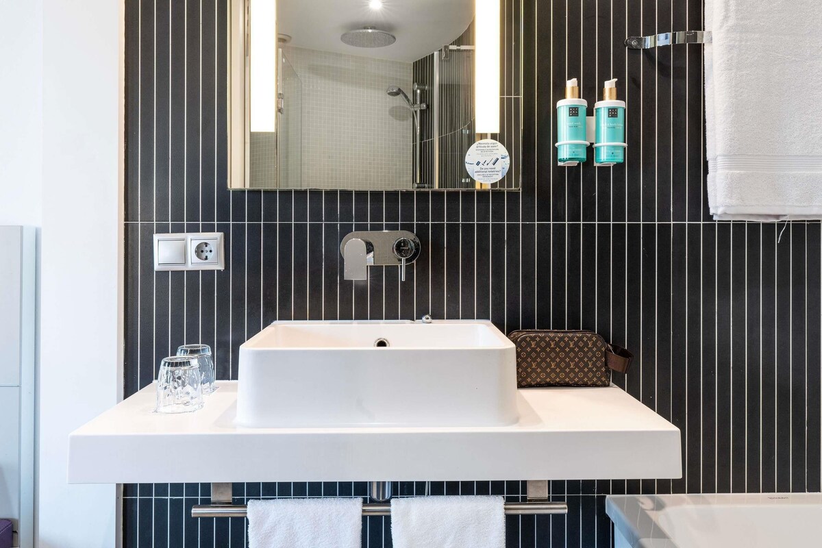 Un baño moderno con un espejo grande, un lavamanos blanco y una ducha. Hay dos botellas de champú y un toallero con toallas.