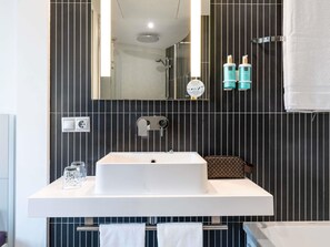 A modern bathroom with a large mirror, a white sink, and a shower. There are two bottles of shampoo and a towel rack with towels. - Novotel Suites Malaga Centro (Málaga)