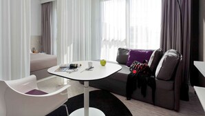 A modern hotel room with a round white table, a grey sofa with purple cushions, and a black floor lamp. - Novotel Suites Malaga Centro (Málaga)
