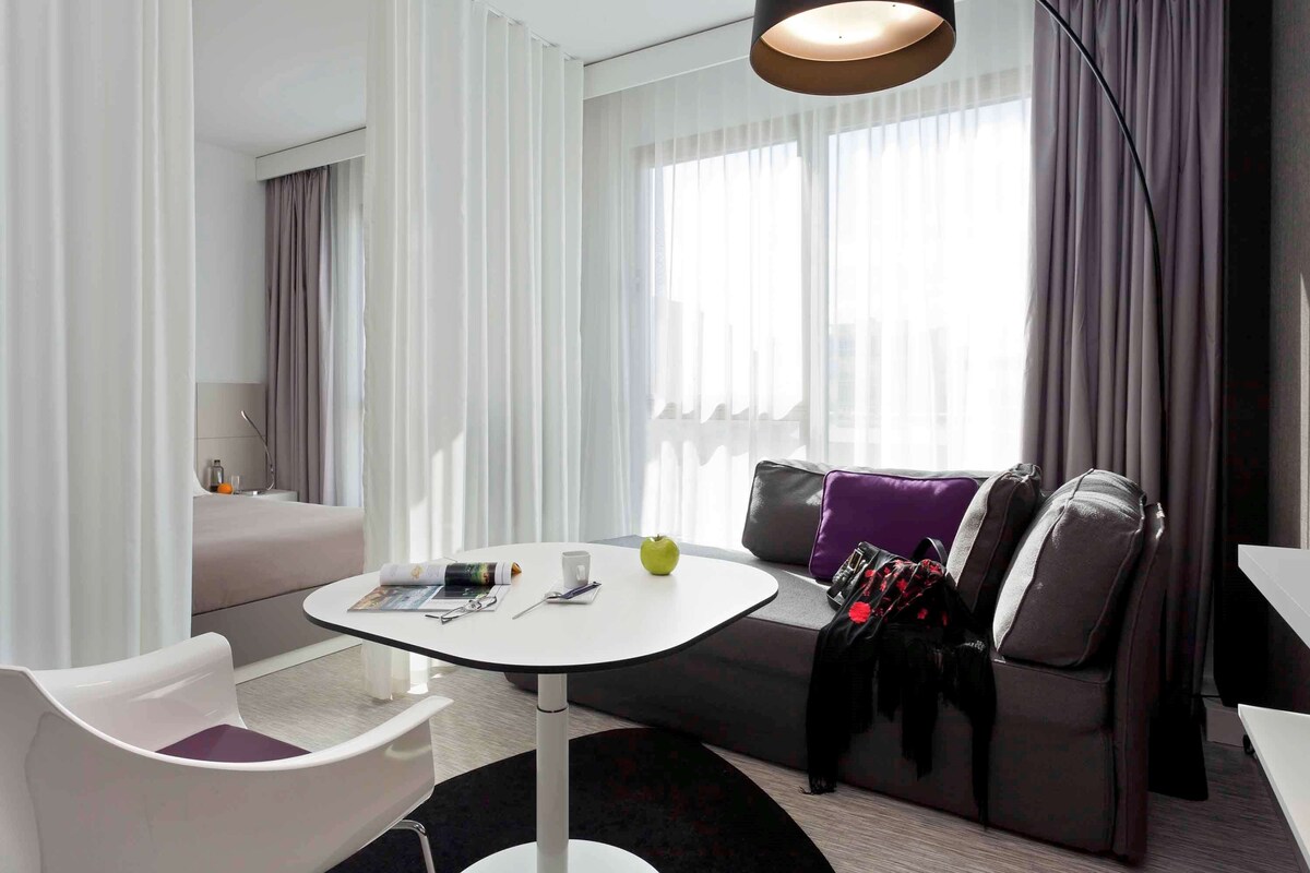 Una habitación de hotel moderna con una mesa redonda blanca, un sofá gris con cojines morados y una lámpara de pie negra.