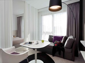 Una habitación de hotel moderna con una mesa redonda blanca, un sofá gris con cojines morados y una lámpara de pie negra.