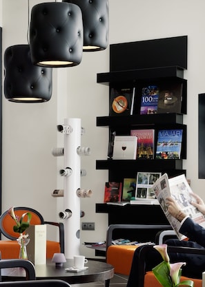 A modern hotel lobby with black tufted chairs, a white column, and a bookshelf with travel books and magazines. - Novotel Suites Malaga Centro (Málaga)