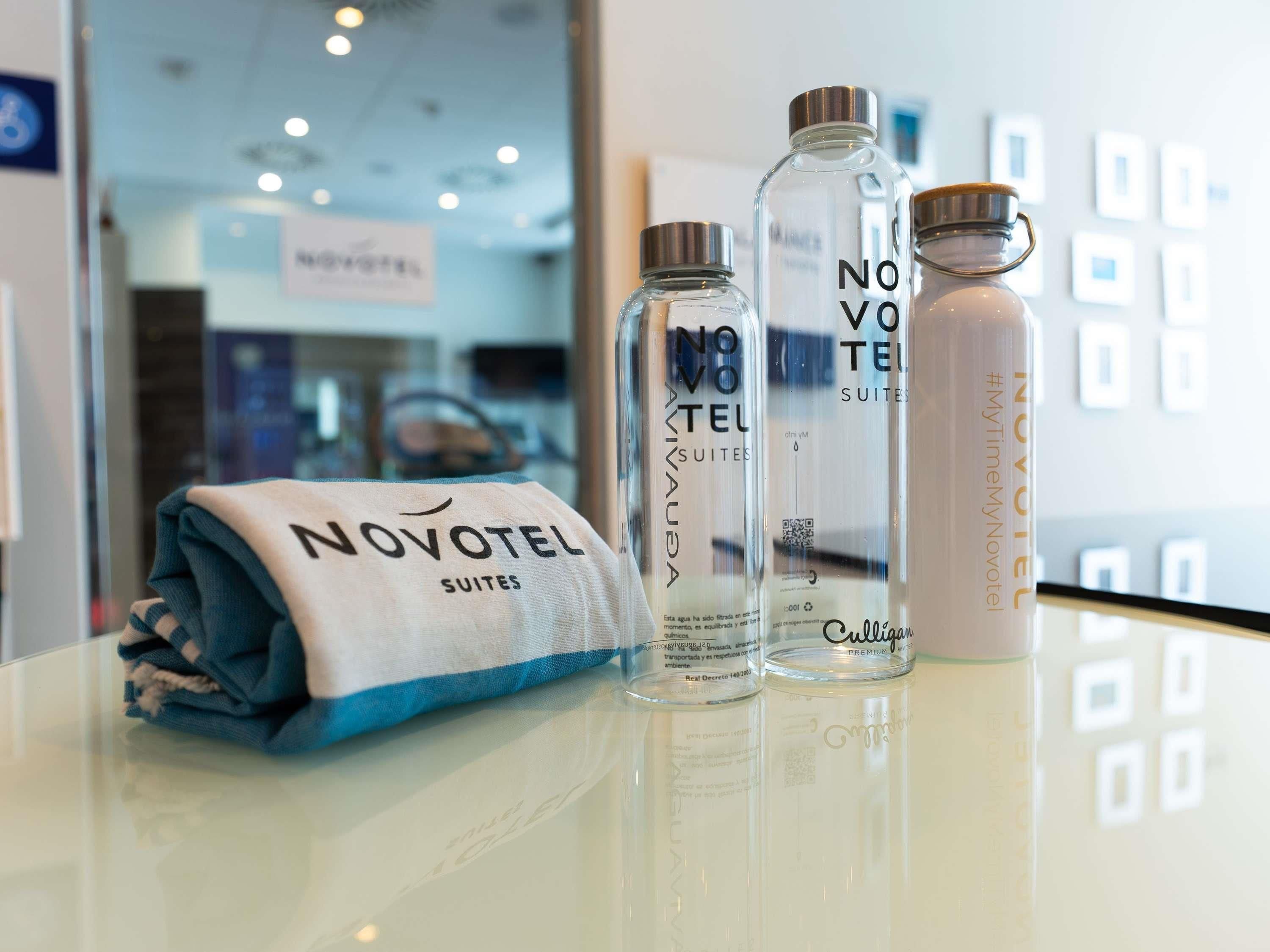 Une chambre d’hôtel avec une serviette pliée avec soin, une bouteille en verre portant l’inscription « NOVOVOTEL SUITES » et une autre bouteille en verre portant l’inscription « CULTURAL PREMIUM ».