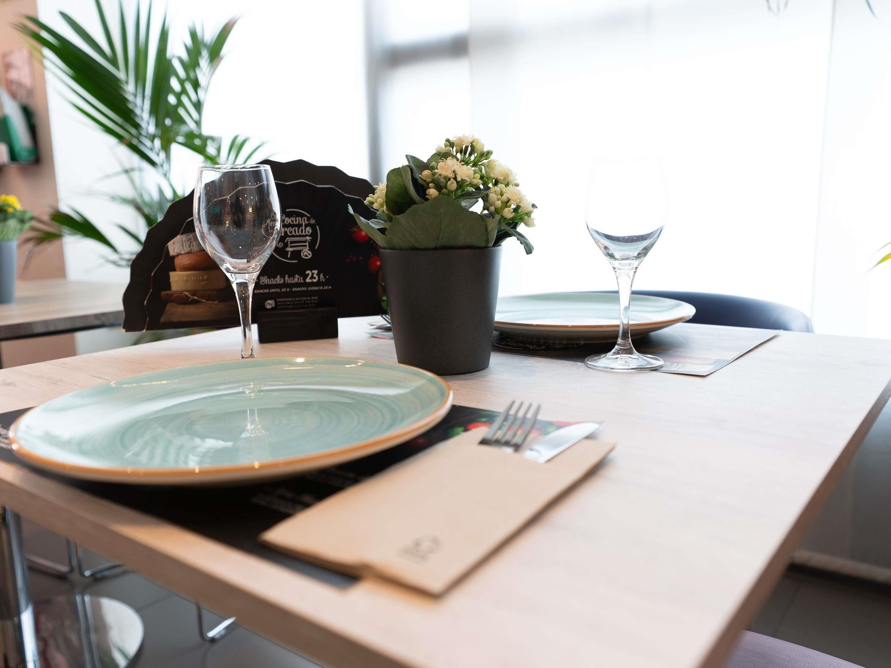 Una mesa dispuesta con platos, cubiertos, una copa, una planta en maceta y un menú.