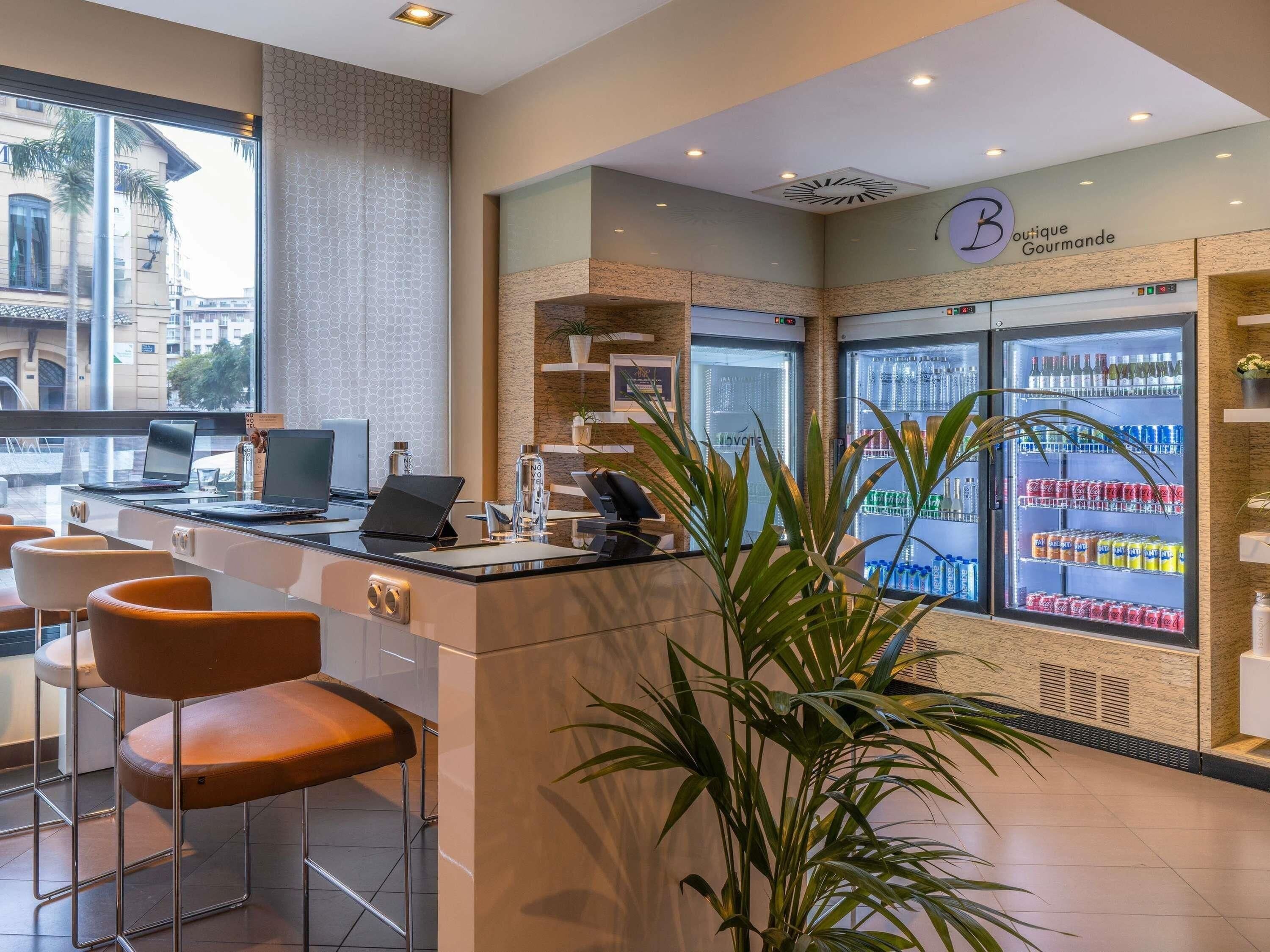 Un área de recepción moderna con mostrador, computadoras portátiles y asientos. Hay refrigeradores llenos de bebidas y un letrero para Boutique Gourmande.