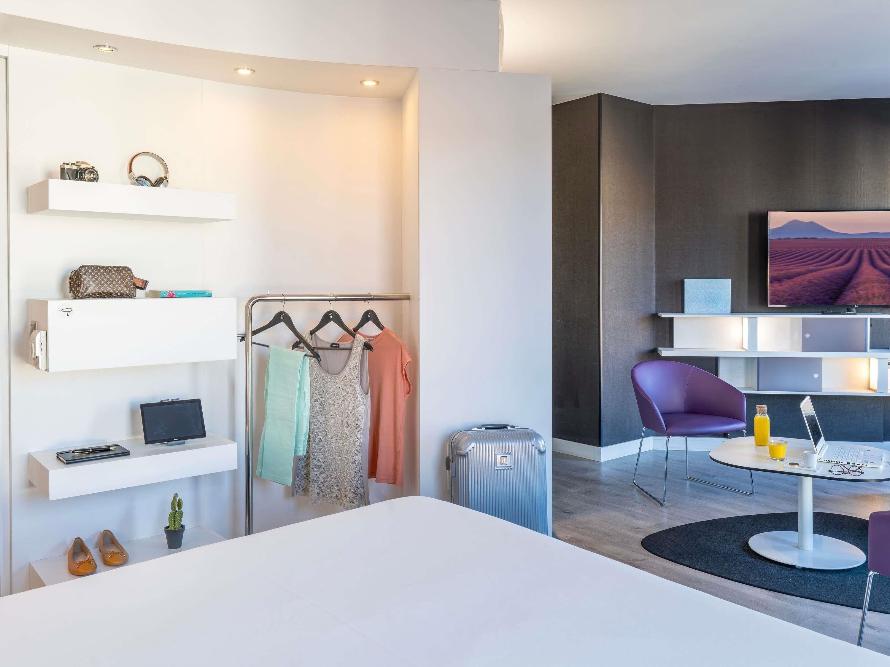 Ein modernes Hotelzimmer mit Bett, Schreibtisch, Laptop, Fernseher und einer Sitzecke mit einem Sessel.