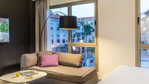 Ett modernt hotellrum med en soffa, ett litet bord och ett stort fönster med utsikt över byggnaderna.