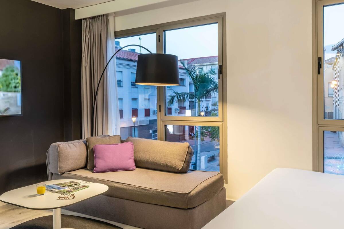 A modern hotel room with a sofa, a small table, and a large window with a view of buildings.