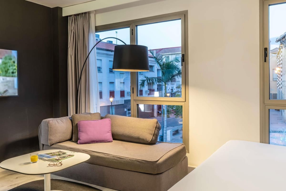 A modern hotel room with a sofa, a small table, and a large window with a view of buildings.