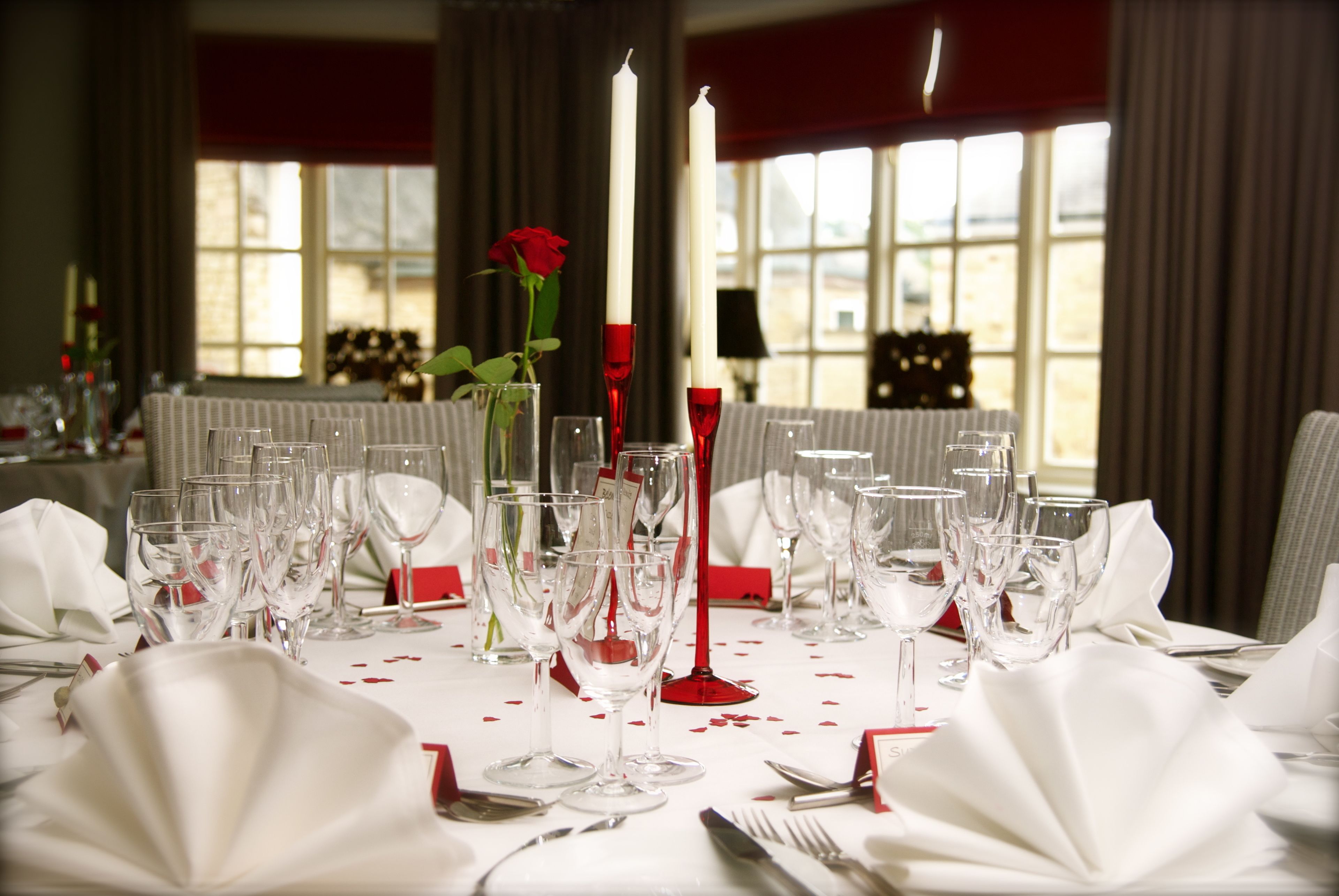 Une table à manger dressée avec plusieurs verres à vin, une rose rouge et des bougies, avec en arrière-plan une fenêtre et des rideaux.