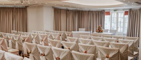 A banquet hall with rows of chairs covered in white fabric and tied with ribbons, a chandelier, and a Christmas tree.