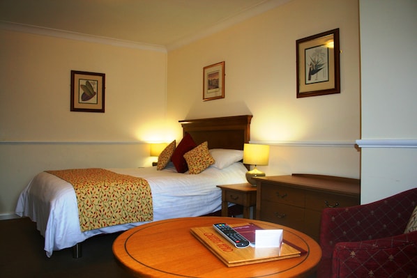 Quarto de hotel com uma cama, abajures, uma mesa de cabeceira de madeira, uma cadeira vermelha e uma mesa redonda com um controle remoto.