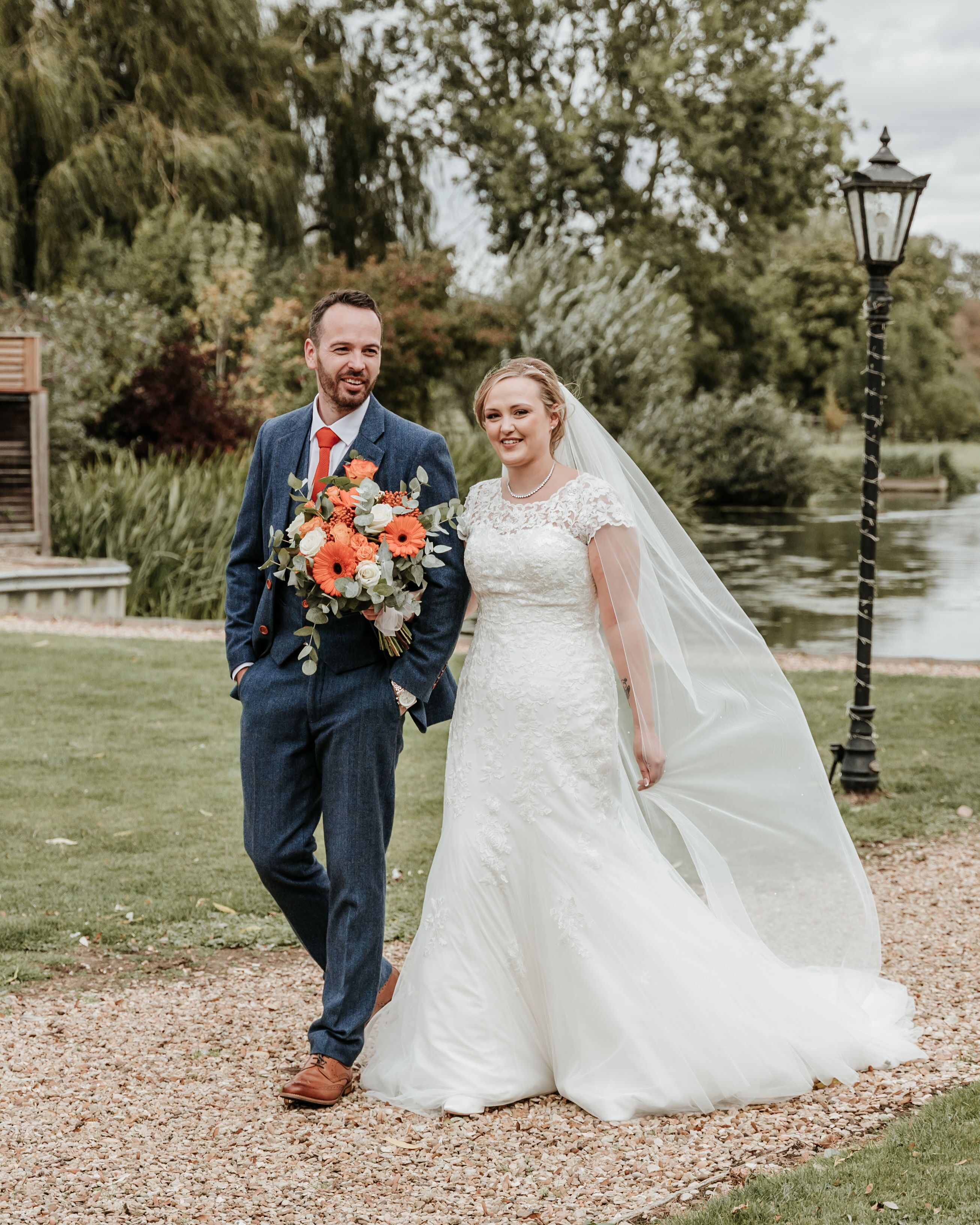 A couple standing on a gravel path with a body of water and greenery in the background.