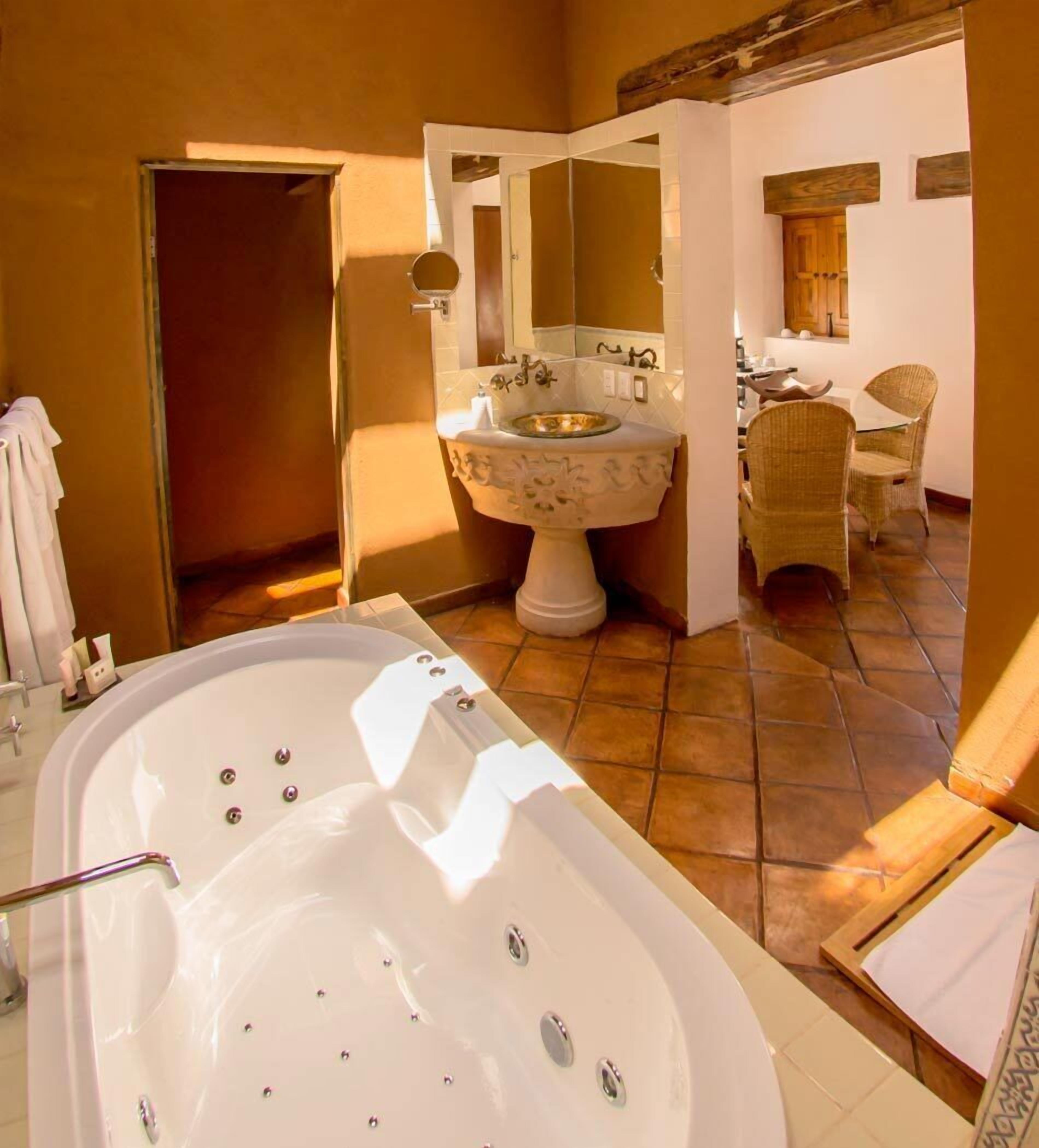 A bathroom with a large bathtub, a pedestal sink, and wicker chairs.