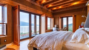 Ein geräumiges Schlafzimmer mit einem großen Bett, einem Schreibtisch und einem Stuhl, mit Holzbalken und großen Fenstern mit Blick ins Freie.