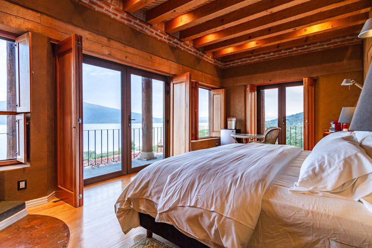 Une chambre spacieuse dotée d’un grand lit, d’un bureau et d’une chaise, avec des poutres en bois et de grandes fenêtres offrant une vue sur l’extérieur.