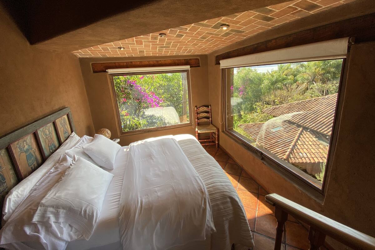 Une chambre avec un grand lit, deux fenêtres et une vue sur la verdure.