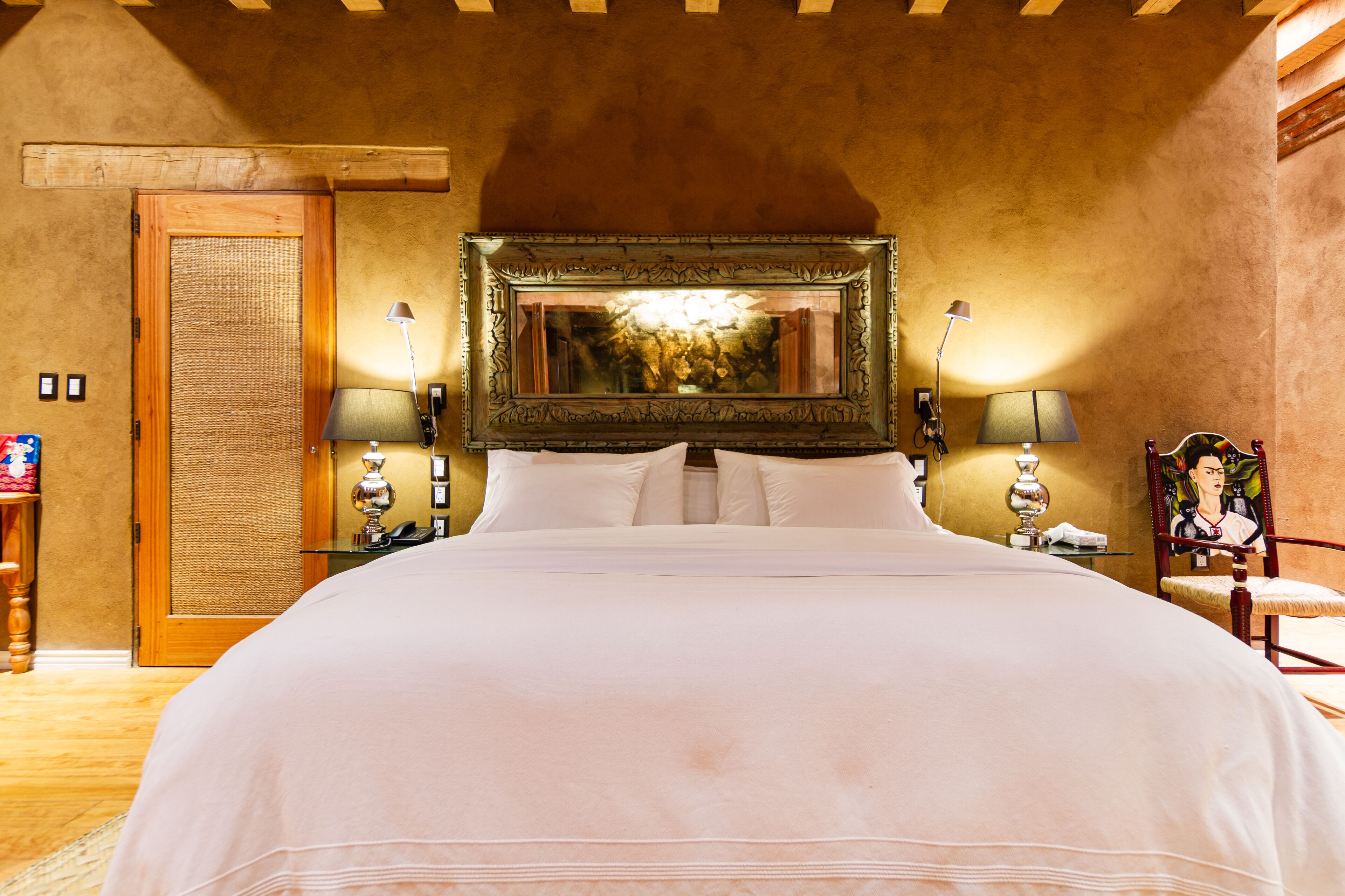 A neatly made bed with a large mirror above it, two bedside lamps, and a framed picture on the wall.