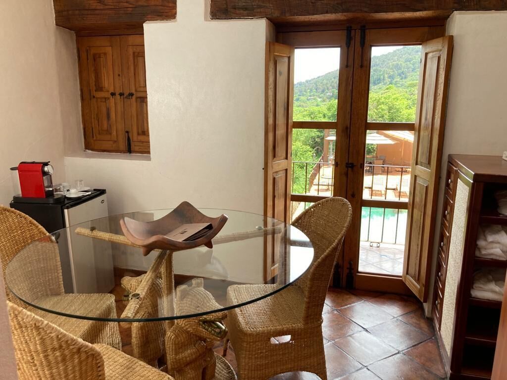 A small dining area with a glass table and wicker chairs, a wooden cabinet, and a view of a balcony with a mountain backdrop.