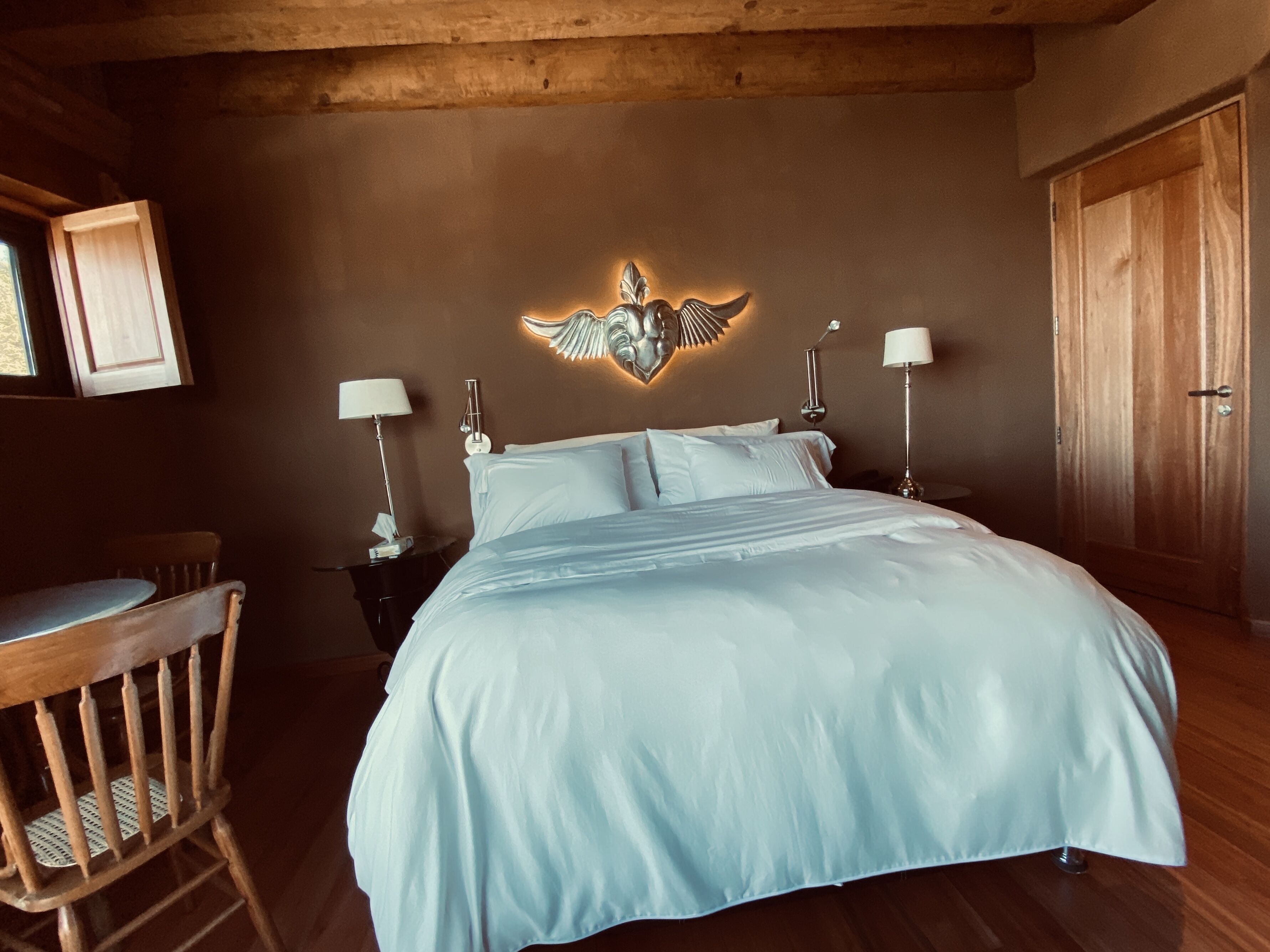 Un dormitorio con una cama grande, dos lámparas de noche, un cabecero de madera, un armario de madera y una mesita pequeña con una silla.