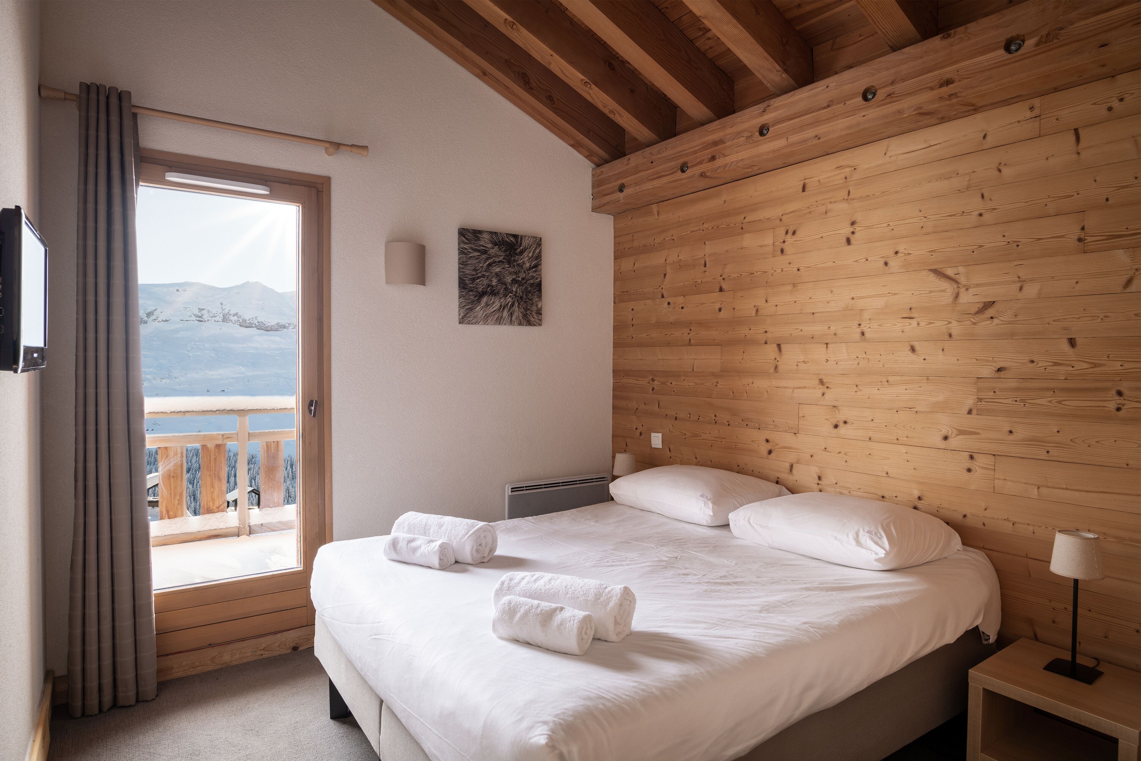 Une chambre avec un plafond en bois, un lit recouvert de draps blancs, une table de chevet avec une lampe, et une vue sur les montagnes par la fenêtre.