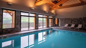 A swimming pool with clear blue water, wooden beams on the ceiling, and large windows offering a view of a wooden deck and greenery. - Dormio Resort Les Portes du Grand Massif (Araches-la-Frasse)