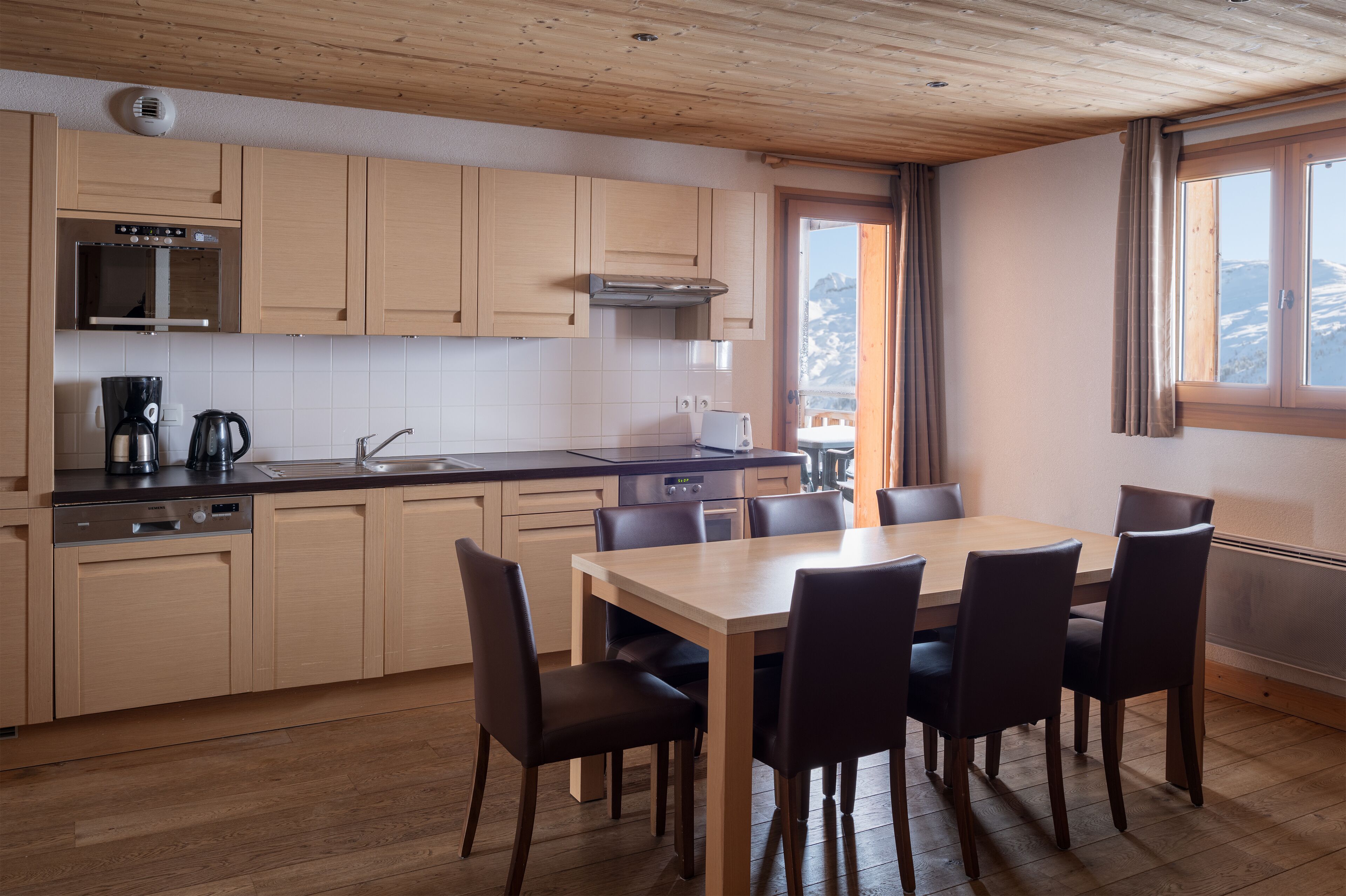 Eine moderne Küche mit Holzschränken, einem Esstisch mit Stühlen und Blick auf Berge durch ein Fenster.