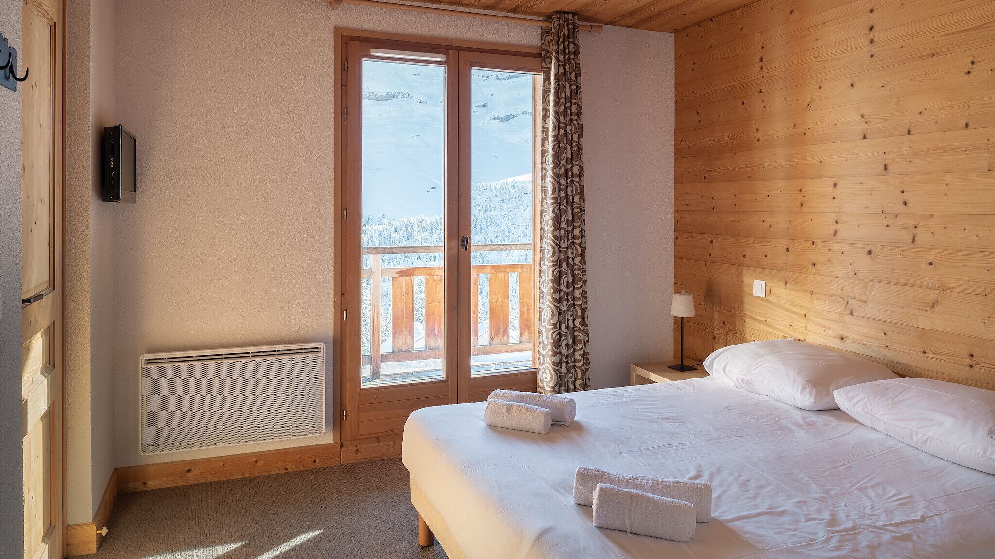 A bedroom with a wooden headboard, a bed with white linens, a nightstand with a lamp, and a view of a snowy mountain through a sliding door.