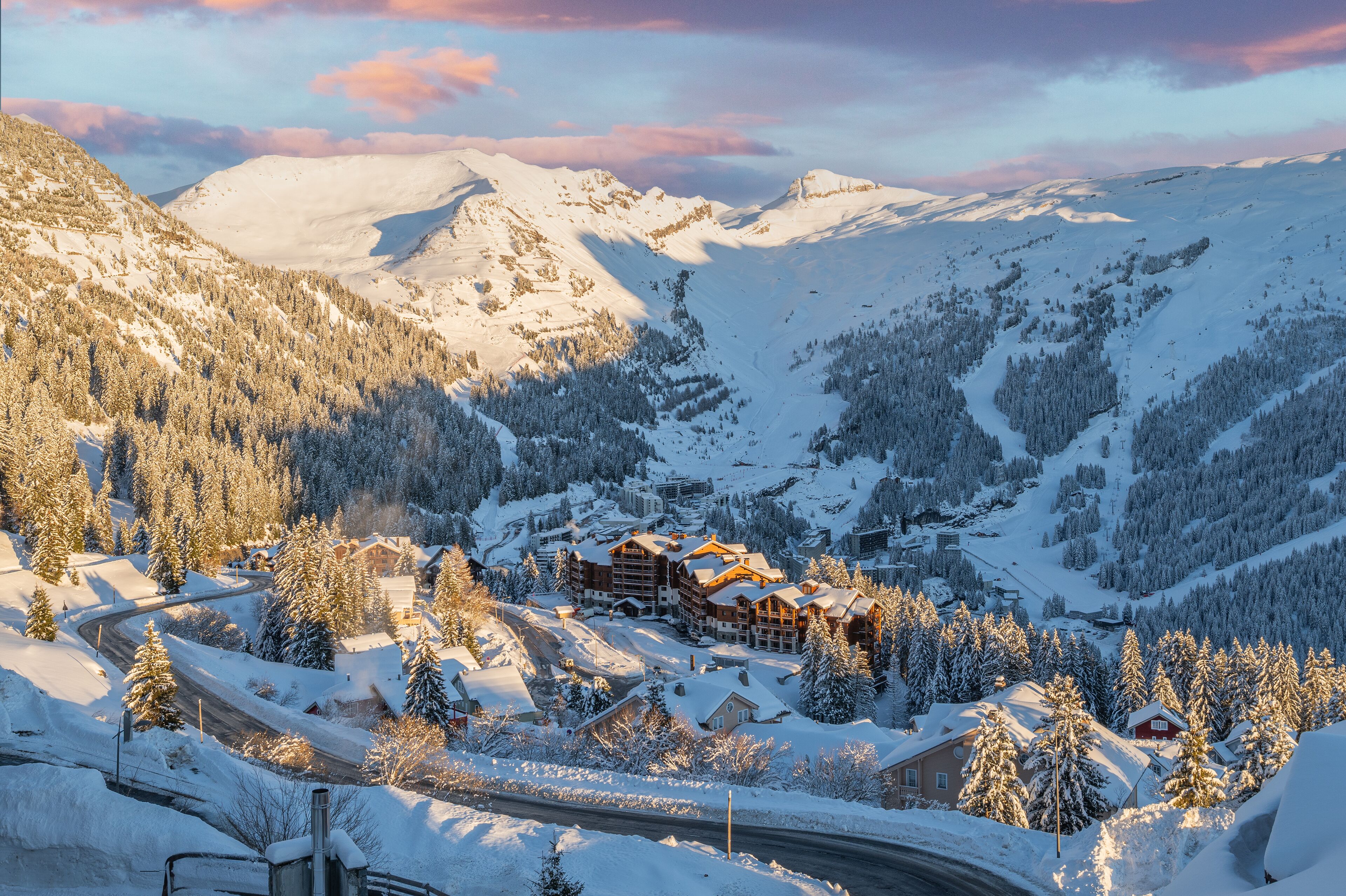 Ein verschneites Bergresort mit mehreren Gebäuden, darunter eine Lodge mit Holzfassade, umgeben von schneebedeckten Bäumen und einer Serpentinenstraße.
