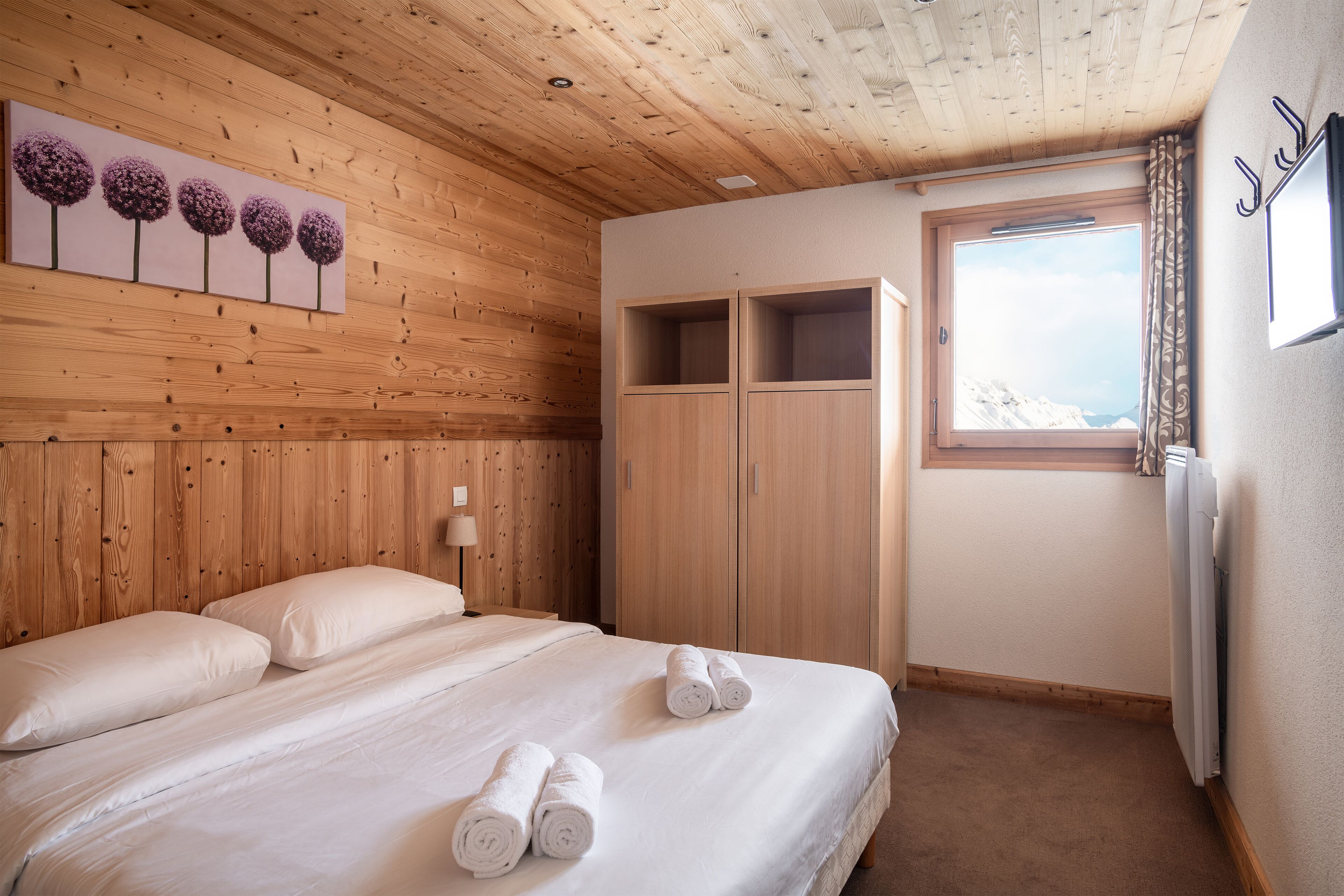Een slaapkamer met een houten plafond, een bed met witte lakens, een schilderij van bloemen, een kledingkast en een raam met uitzicht.