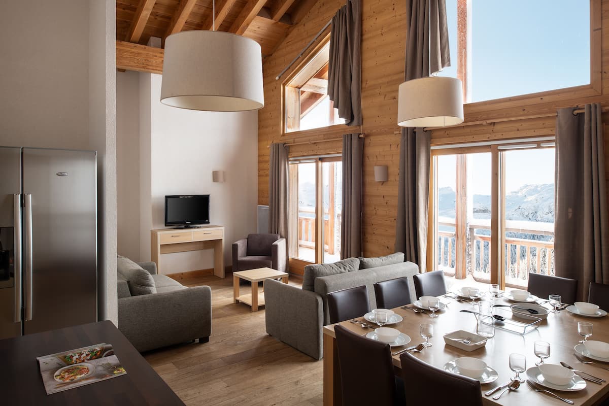 A modern, well-lit living room with a dining area, large windows, and a television.