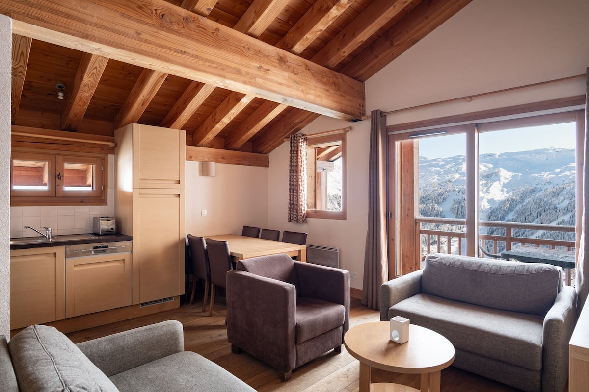 A cozy cabin with wooden beams, a kitchen with a dining area, and a large window offering a mountain view.