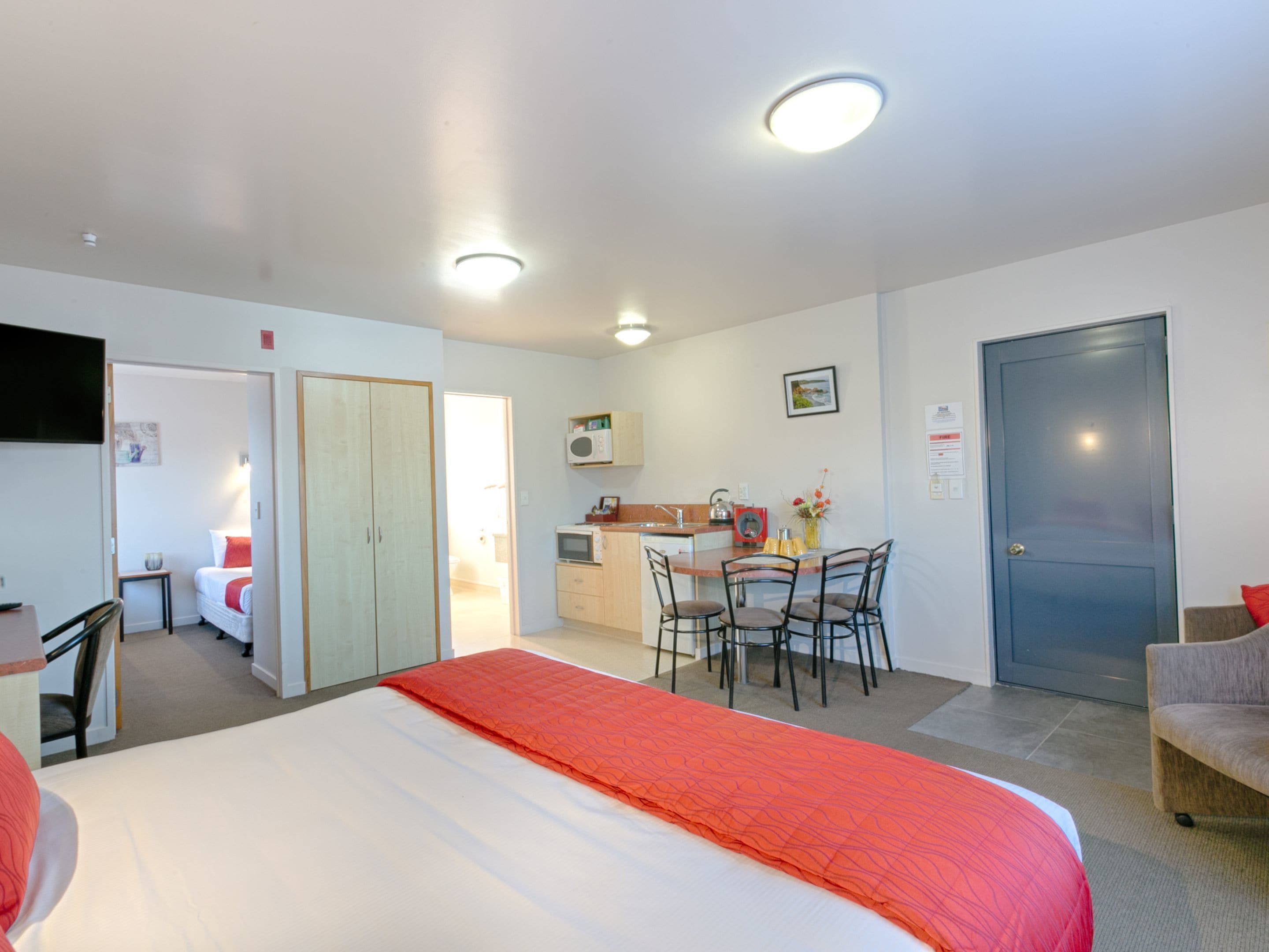 A hotel room with a bed, a desk, a chair, a kitchenette, and a door.
