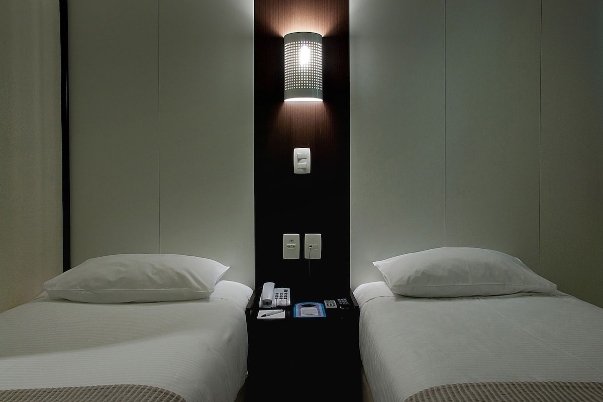 A hotel room with two single beds, a nightstand with a phone and remote control, and a wall-mounted light fixture.