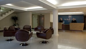 Un área de recepción de hotel con diseño moderno, que incluye un mostrador de recepción, varias sillas de cuero, una escalera y una planta grande en maceta.