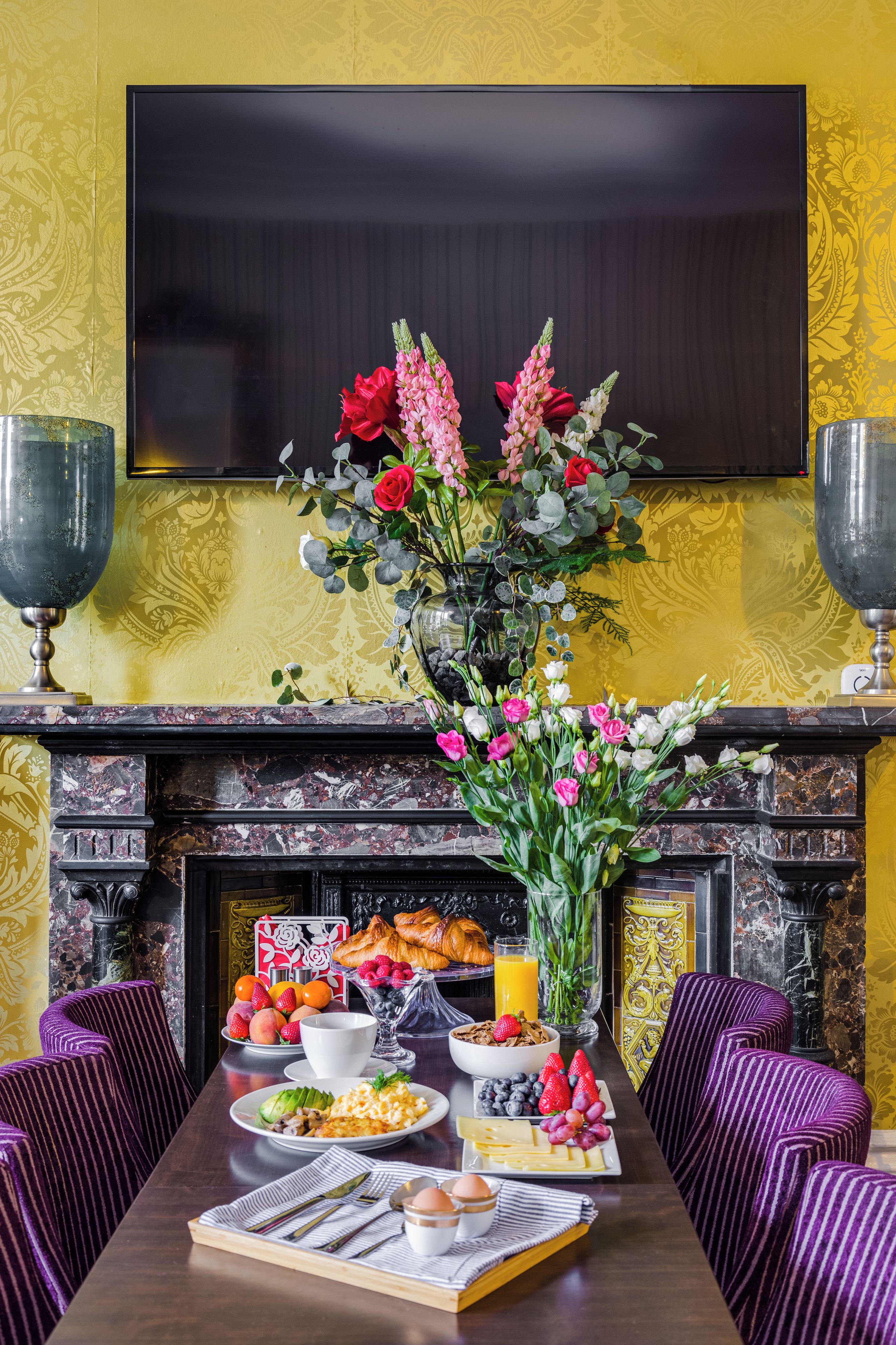 A dining table set with breakfast items, a vase of flowers, and a fireplace.