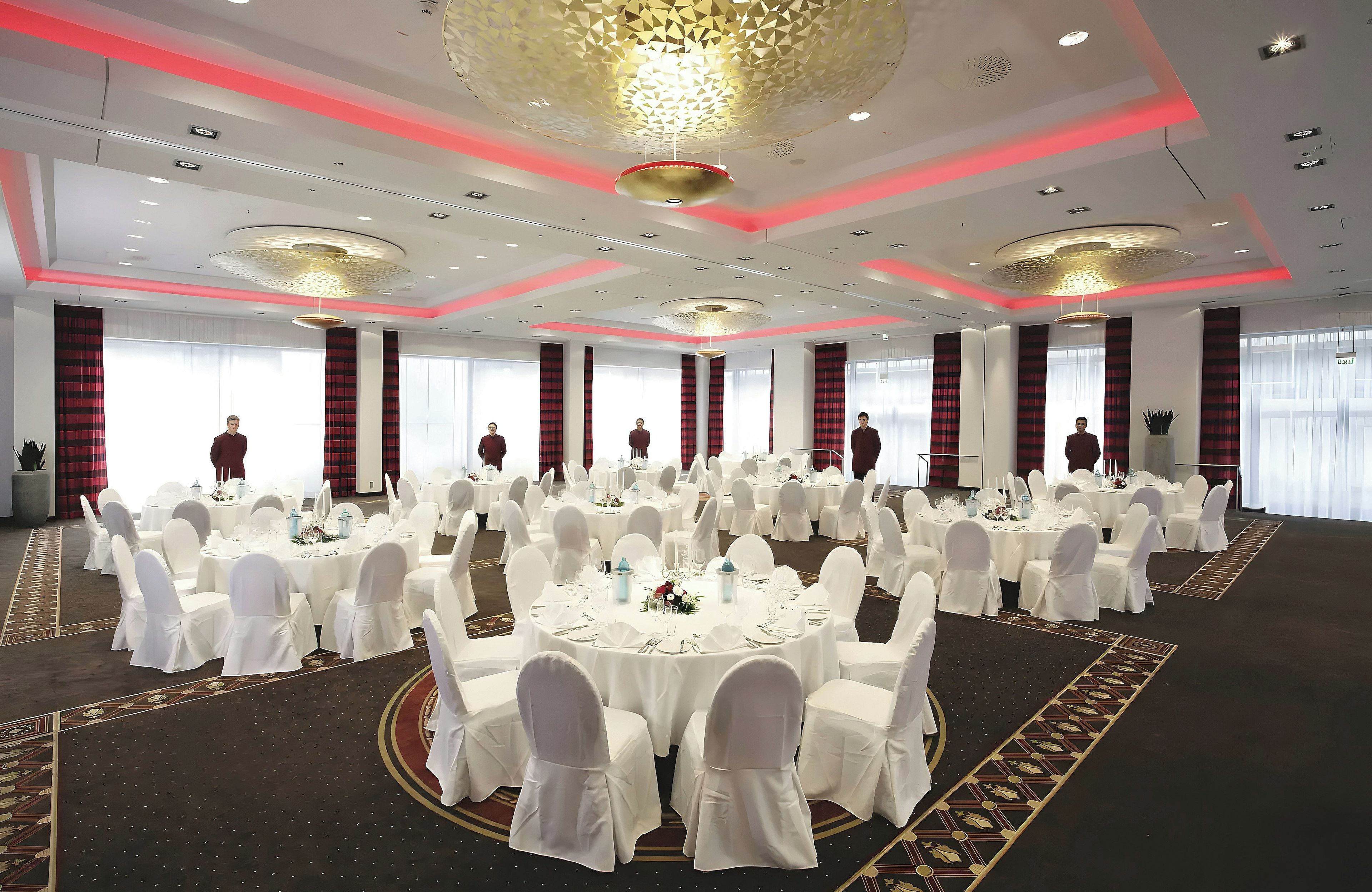 A banquet hall with round tables, white chairs, and red curtains.