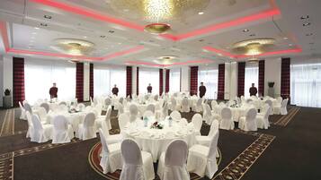 Un salón de banquetes con mesas redondas, sillas blancas y cortinas rojas.