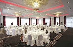 Un salón de banquetes con mesas redondas, sillas blancas y cortinas rojas.