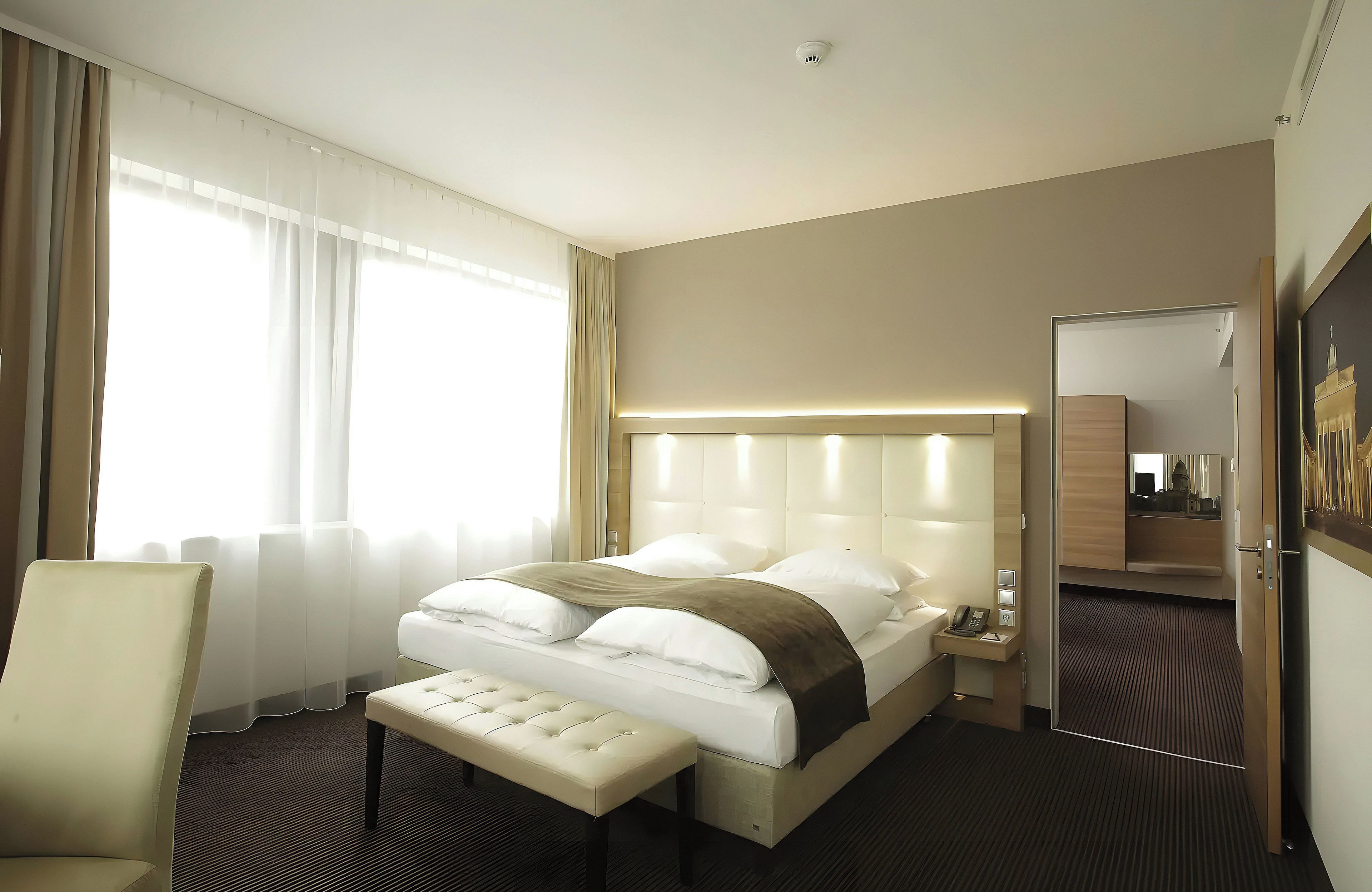 Ein Hotelzimmer mit einem großen Bett, einem Nachttisch mit Telefon, einem Sessel und einem Fenster mit Vorhängen.